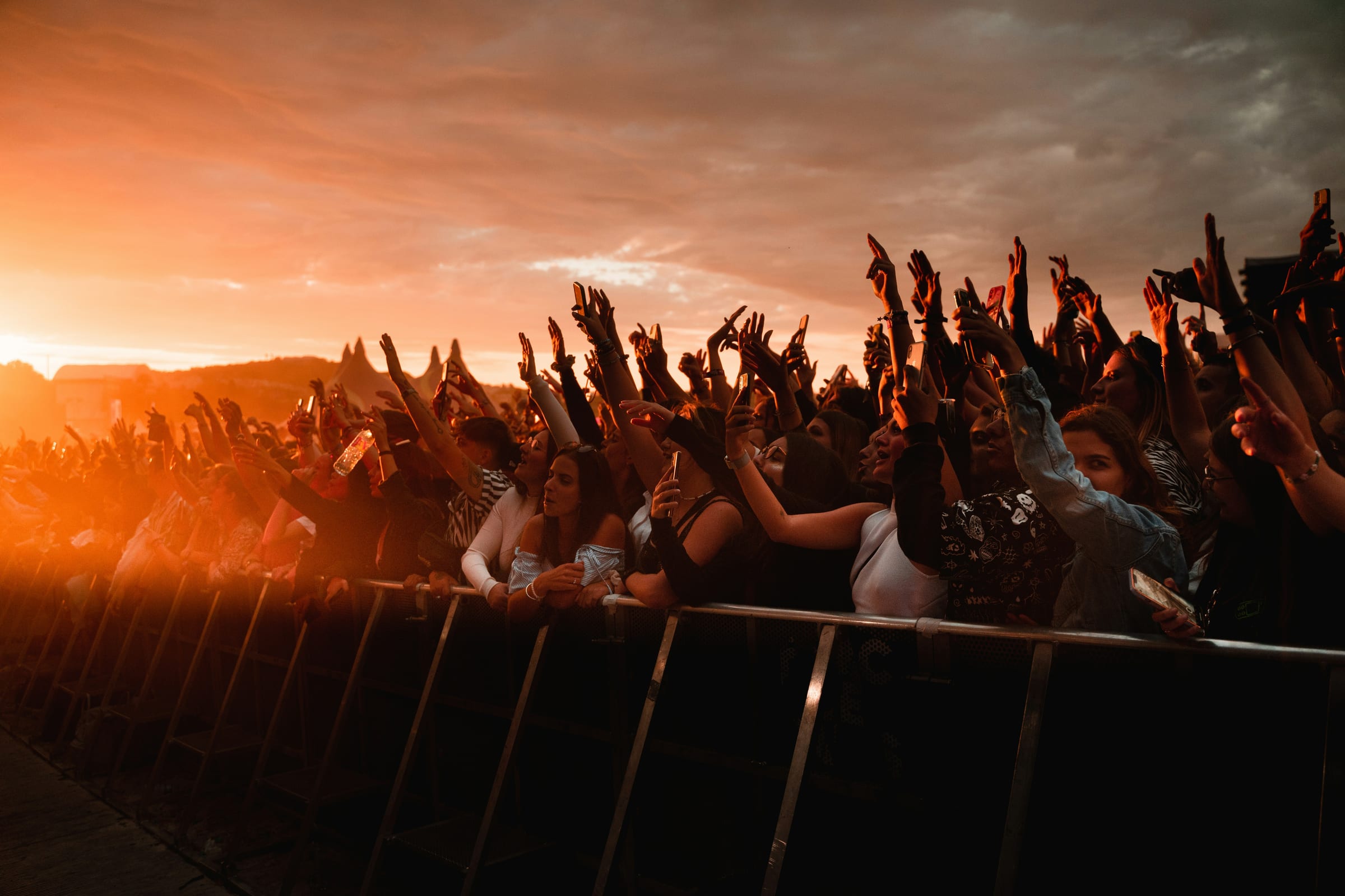 A large crowd at an outdoor festival with hands raised, lit by a warm sunset.