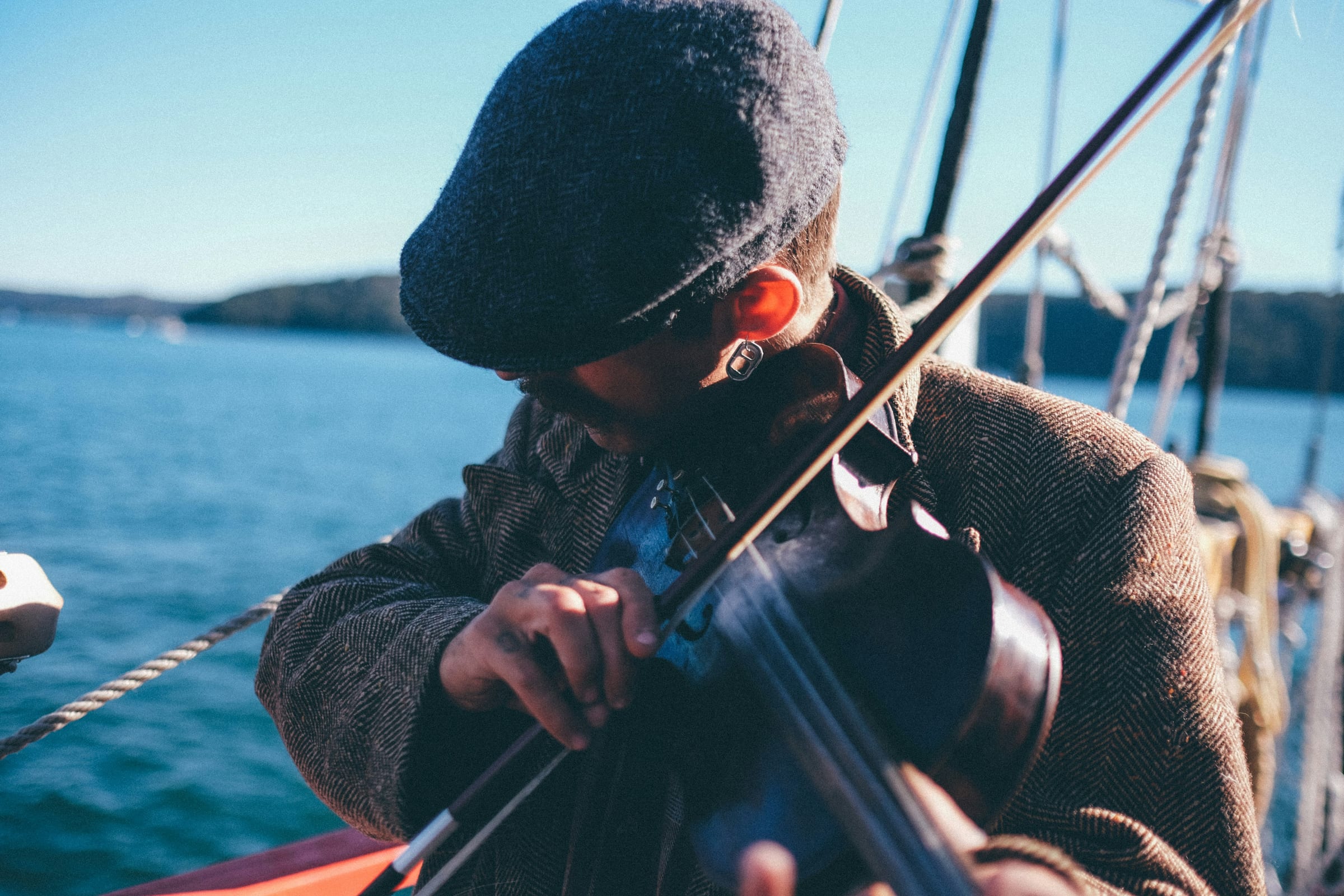 A musician in a flat cap playing a violin on the deck of a boat by the coast.